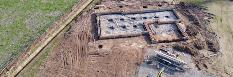 Photo shows a brown field with heavy machinery construction some foundations for a building.